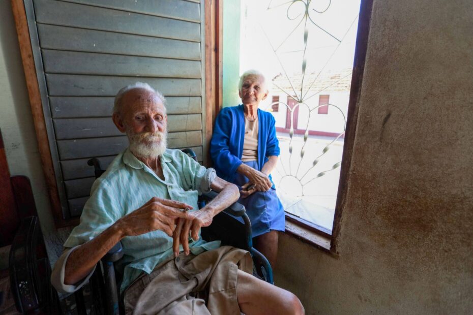 Idoso em cadeira de rodas ao lado de uma idosa em casa